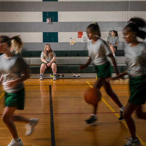 High school girls playing basketball in a gym as a heavy set girl sits in the background watching
